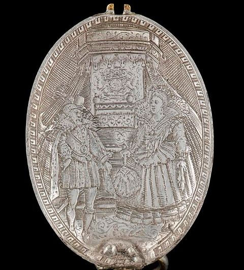 Photograph. An oval, silver object is photographed against a black background. A decorative geometric pattern surrounds the edge of the object. Within this frame an illustration features a man and a woman standing side by side in historical clothing – he wears a fitted tunic and she wears a long dress with a corset waist. Behind them is a tall stone structure with a hexagonal shape.