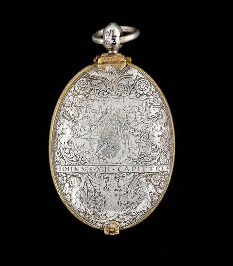 Colour photograph. Documentation of a silver, oval object photographed against a black background. The object has a gold trim around the edge, a clasp visible at the base, and a metal loop at the top. Its surface is inscribed with an ornate pattern.