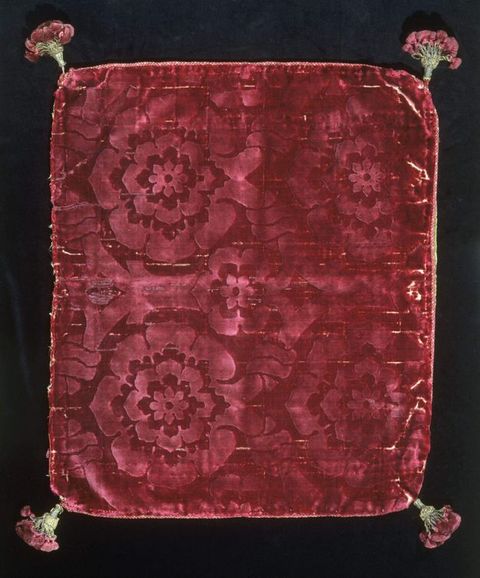 Fabric cushion. A document of a dark red, velvet cushion cover photographed against a black background. The blanket features a large, yet subtle floral pattern, and has green and red tassels in each corner.