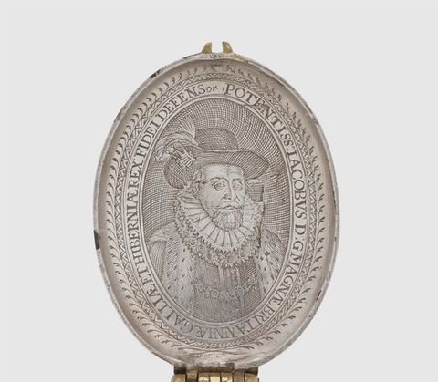 Colour photograph. The upper, inner lid of an oval, sliver object is photographed against a white background. It is decorated with an embossed portrait of a man wearing a fur-trimmed cape, a stiff, ruffled collar, and a hat with a feather. Text encircles the inner rim of the oval. A hinge connecting it to the lower half is just visible at the bottom. 