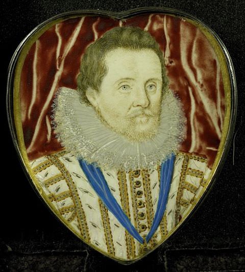 Colour photograph. A colour image showing a heart-shaped miniature portrait of a man’s head and shoulders in historical dress, wearing a white jacket with gold details, and a stiff white circular collar. A blue sash is hung around his neck. Red, draped fabric can be partially seen behind him.