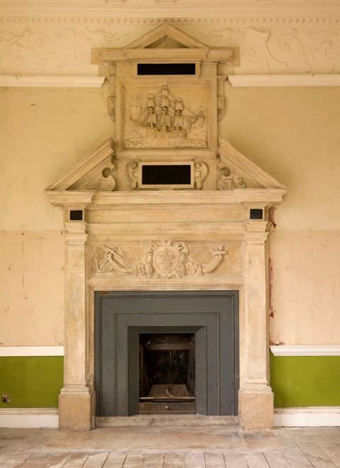 Colour photograph. A document of a historical interior, showing an ornate fireplace with detailed sections that extend upwards towards the ceiling. In the surrounding room are pale wood floorboards, a detailed cornice, and green sections of paint on the lower walls.